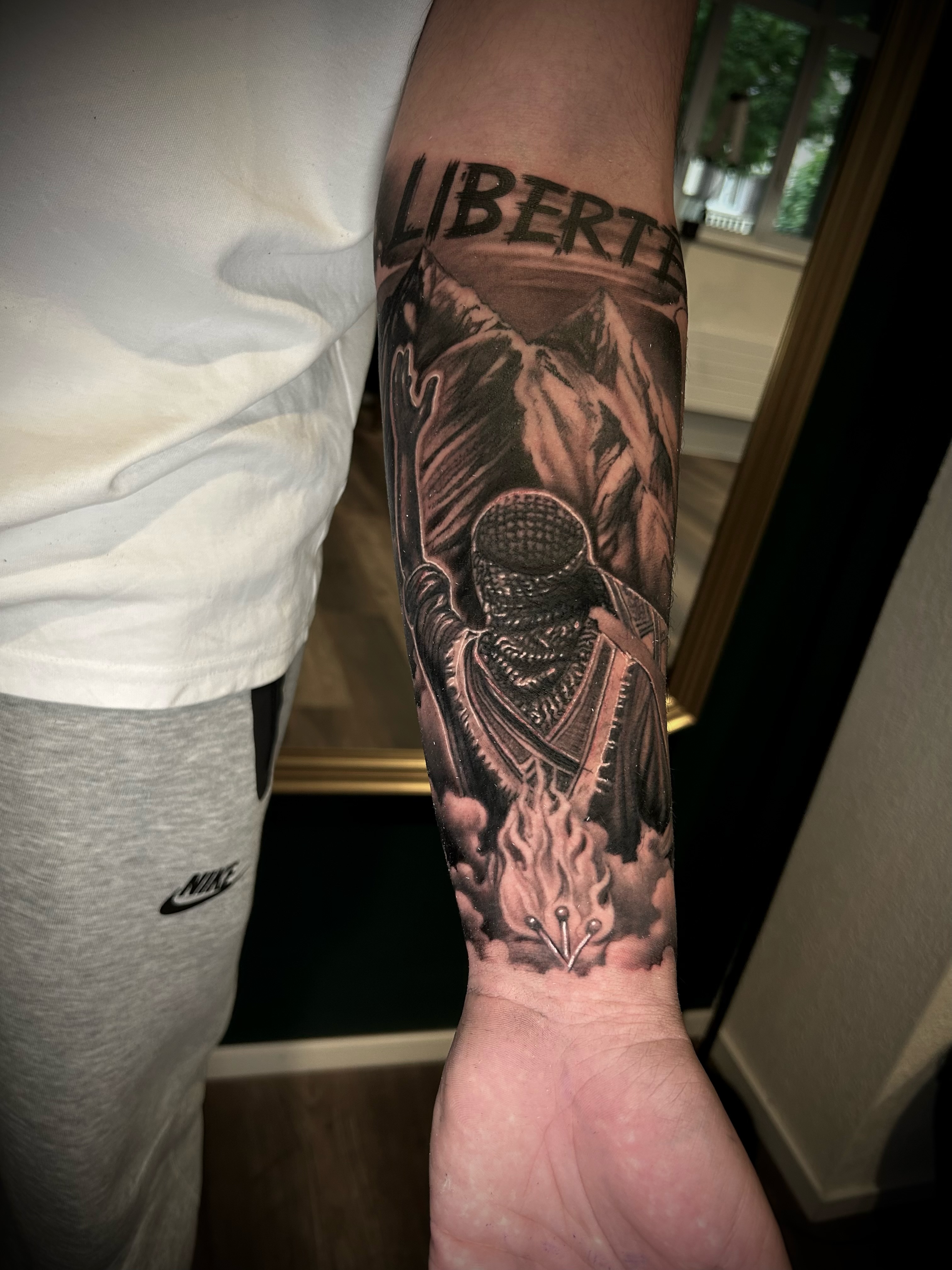 Lettering-Tattoo Liberté mit Bergpanorama und Feuer am Unterarm